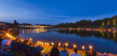 Menschen sitzen mit Picknickdecken um den Hollersee und lauschen einem klassischen Konzert 